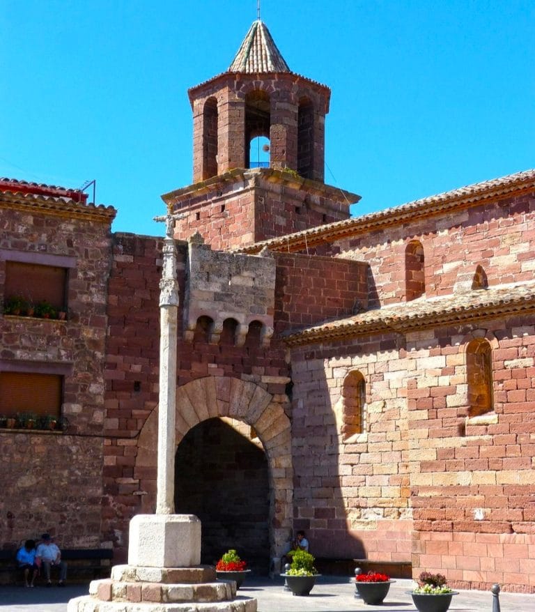 Plaza Mayor de Prades con iglesia de piedra rojiza y cruz medieval, en el corazón del pueblo histórico del Baix Camp, Tarragona.