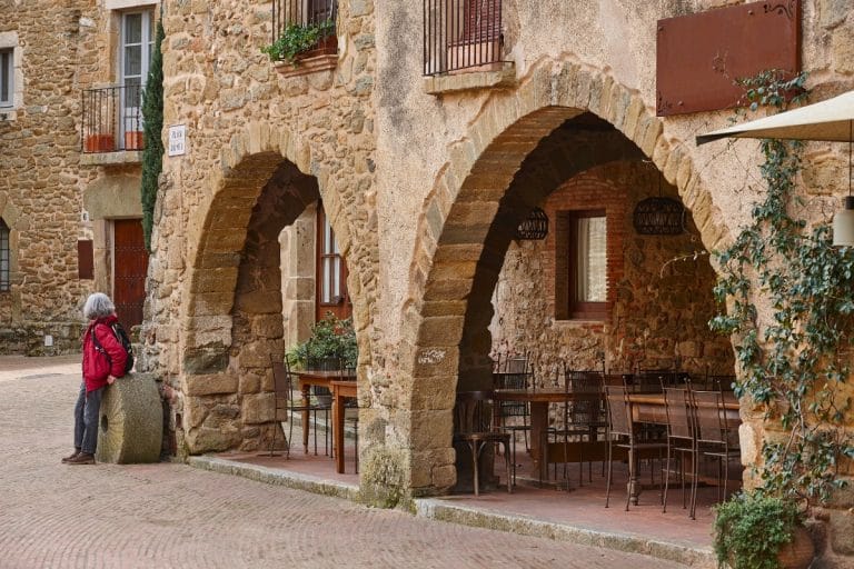 Porches de piedra con mesas y arcos en la Plaça Jaume I de Monells, Girona.