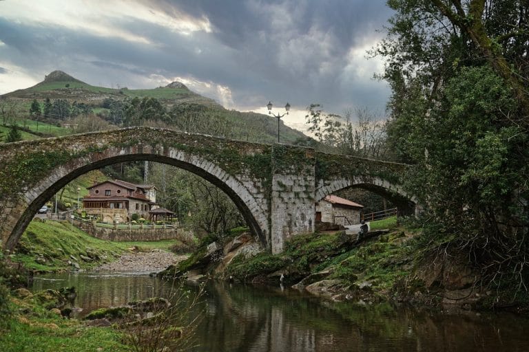 Puente medieval en Cantabria sobre un río, con casas rurales al fondo y montañas verdes