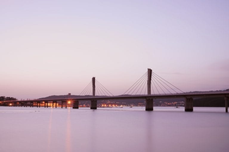 Puente sobre la ría de Noia al atardecer con luces urbanas al fondo y cielo en tonos pastel