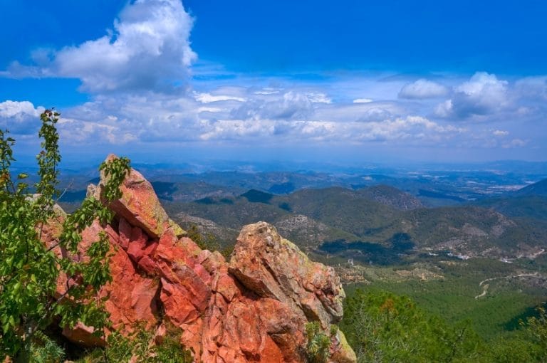 Vista panorámica de la Sierra de Espadán, mostrando sus características formaciones rocosas de tonos rojizos y verdes, bosques frondosos y un cielo azul con nubes.
