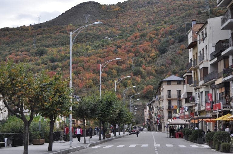 Calle principal de Sort en Lleida con terrazas y árboles en tonos otoñales frente a una ladera de montaña