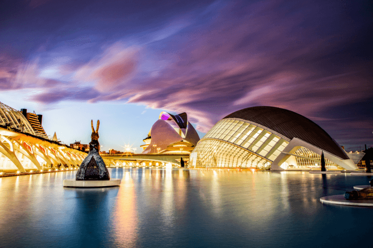 Vista al atardecer de la Ciudad de las Artes y las Ciencias en Valencia, con sus edificios de arquitectura moderna iluminados reflejándose en el agua y un cielo púrpura.