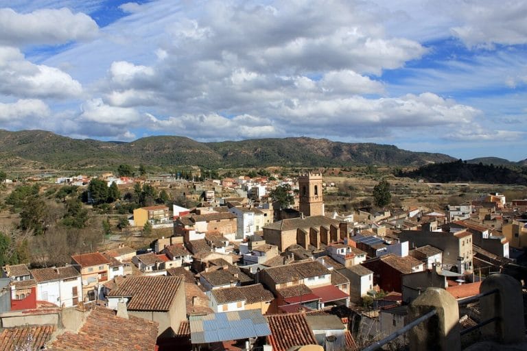 Vista panorámica de un pueblo valenciano rodeado de montañas, con calles estrechas y tejados tradicionales bajo un cielo parcialmente nublado