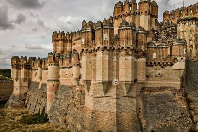 Vista exterior del Castillo de Coca, mostrando la elaborada arquitectura mudéjar de ladrillo con numerosas torres y un foso seco en la parte inferior, bajo un cielo nublado