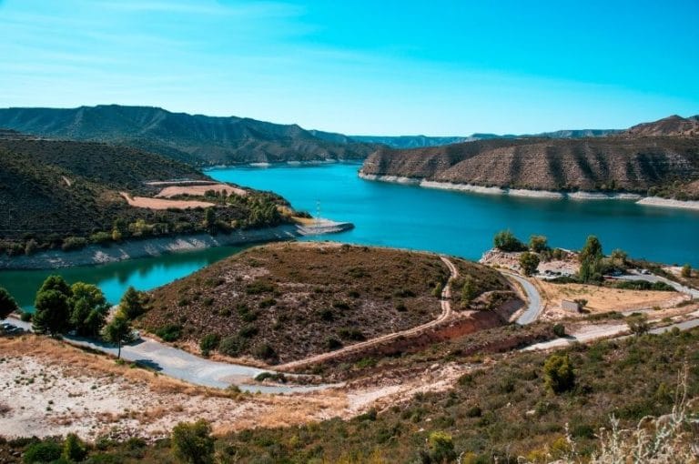 Vista panorámica del embalse de Mequinenza, mostrando sus aguas azules rodeadas de colinas áridas y formaciones rocosas, con una carretera sinuosa en la orilla.