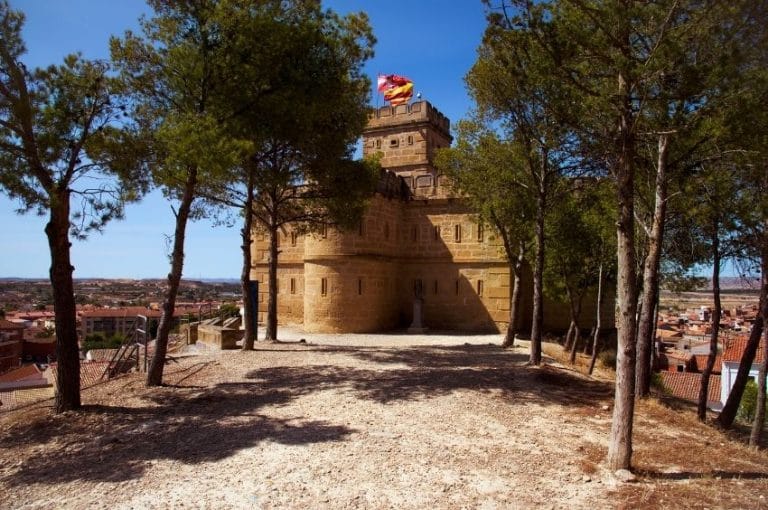 Vista de la Torre de Salamanca en Caspe, un edificio de piedra con varias ventanas y una bandera de España en lo alto, rodeado de pinos y con vistas a un pueblo.
