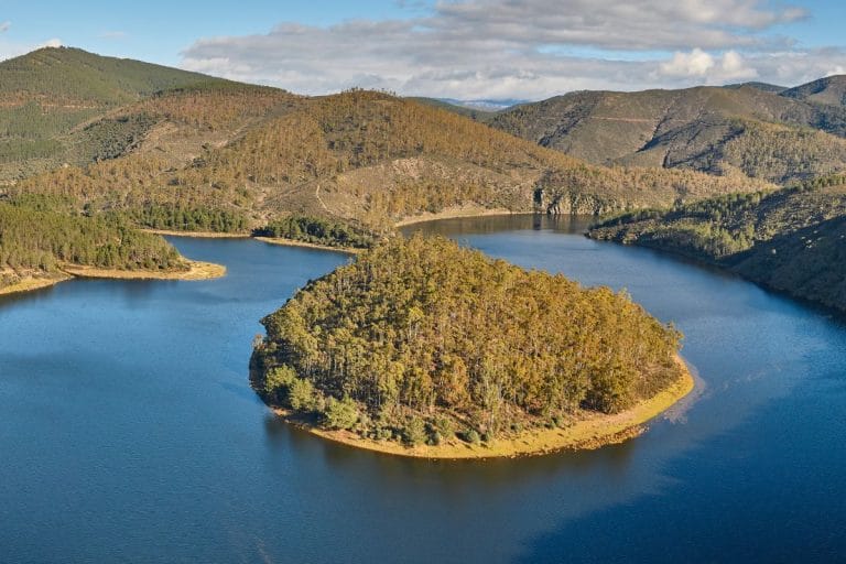 Vista aérea del meandro del Melero, destacando una formación de tierra en forma de isla con vegetación frondosa en el centro de las aguas azules del río, rodeado de colinas boscosas bajo un cielo parcialmente nublado