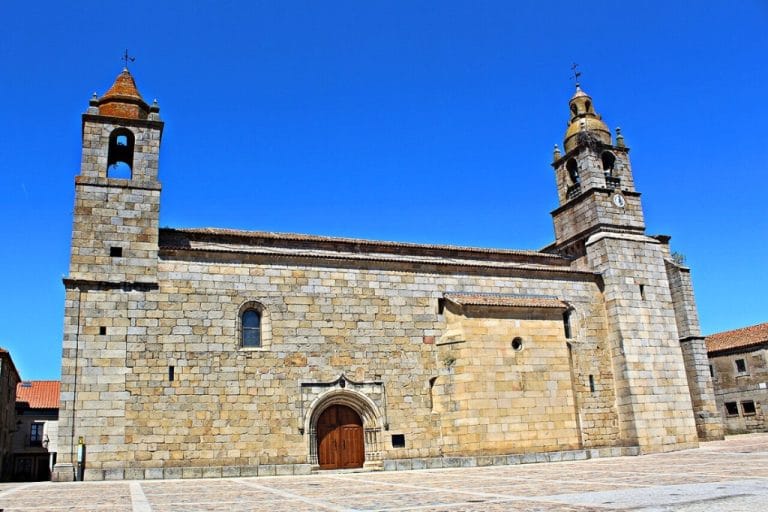 Vista de la fachada principal de una iglesia de piedra en San Felices de Gallegos, con dos torres y un campanario, en una plaza empedrada bajo un cielo azul.