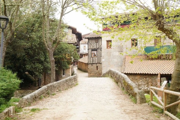 Calle empedrada de San Martín del Castañar, con un pequeño puente de piedra que cruza un arroyo y casas tradicionales de arquitectura de piedra y madera.